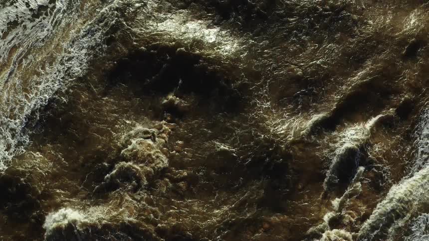 An intense aerial shot captures heavily churned dark muddy brown water large active waves break diagonally across the frame producing significant patches of thick white foam