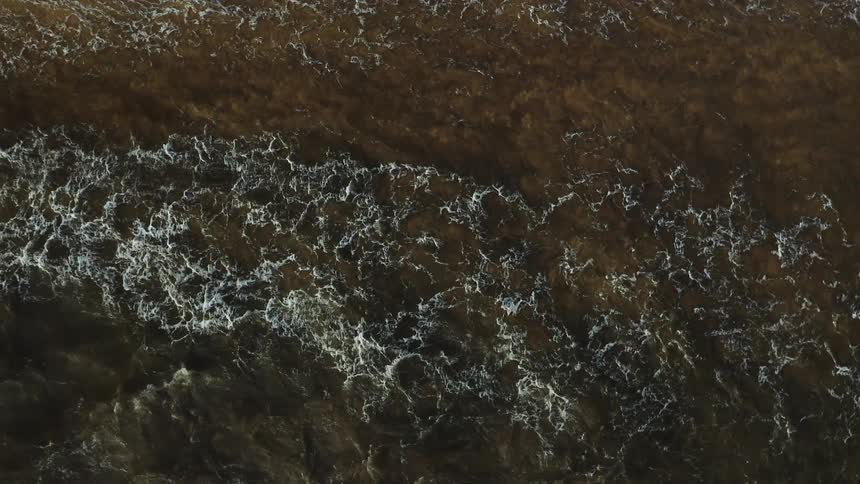 Aerial view of choppy muddy brown ocean water with white foam patterns the camera looks straight down as waves move across the frame with a bird briefly flying into view