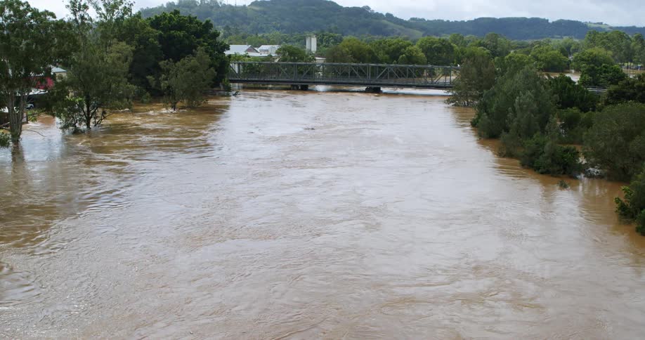 An elevated wide shot of a brown fastflowing river during a flood event with the high water level submerging the adjacent riverbanks and flowing under a metal truss bridge in the background