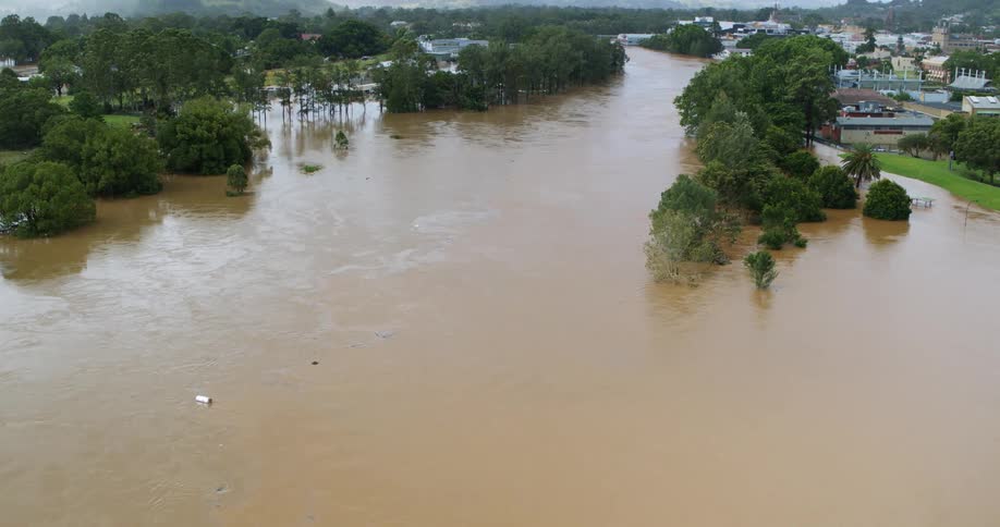 An aerial view captures a powerful brown muddy river overflowing its banks due to significant flooding submerging trees and approaching the edge of an adjacent highway overpass on the right