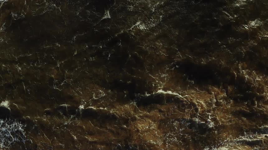 A close aerial shot looks down onto dark agitated and murky brown water the turbulent surface is streaked with white foam where waves are actively breaking
