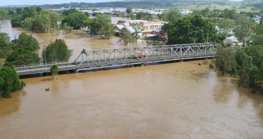 An aerial shot shows a muddy overflowing river during a flood with the water inundating adjacent land and buildings an orange and white emergency vehicle crosses a lowlying steel truss bridge in the center frame