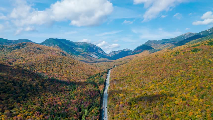 Long road cuts through a mountain valley covered by forest in autumn colors under a partly cloudy sky