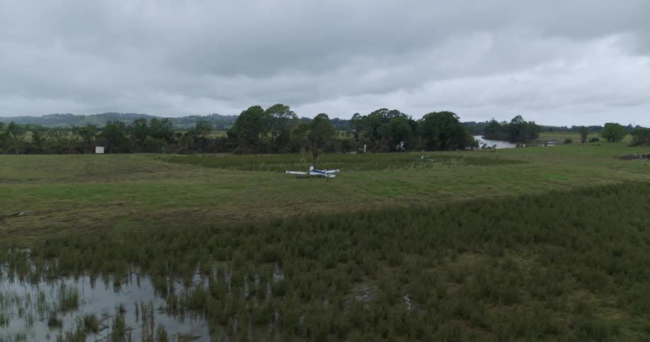 Small aircraft in grassy field surrounded by wetland vegetation and cloudy sky in rural area