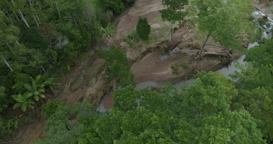 An aerial view shows a river cutting through dense green forest with a large exposed muddy bank and uprooted trees revealing the force of the water