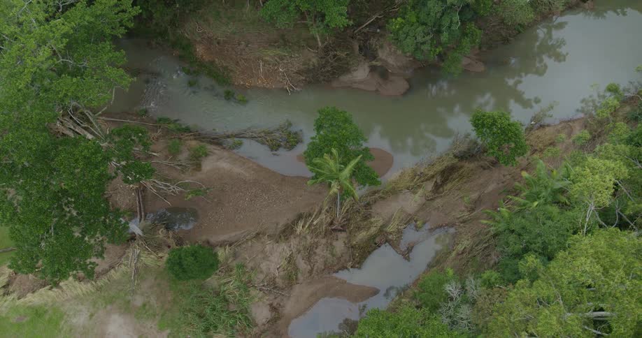 Muddy watercourse winds through a rural area with eroded banks trees and pools formed by flooding