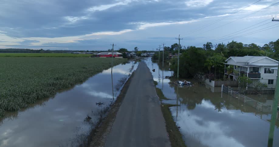 A quiet flooded rural road reflects the cloudy sky with houses and fields submerged under calm murky water