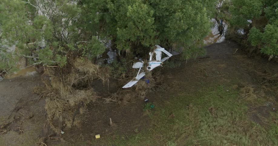 An aerial shot captures the wreckage of a small white airplane lying broken on the ground amidst trees and a large accumulation of natural debris in a muddy rural area
