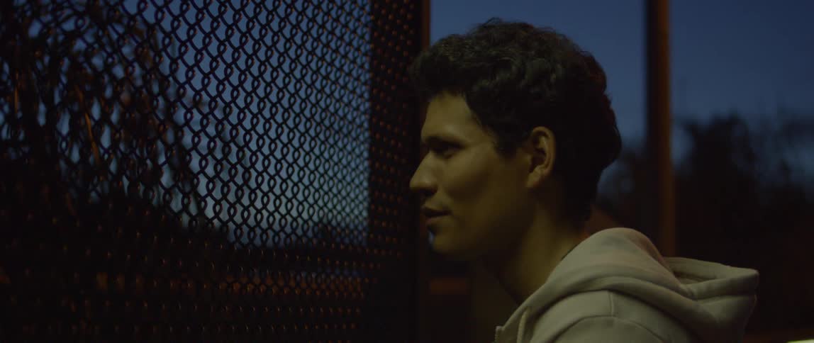 Man stars through a chain link fence outside at night