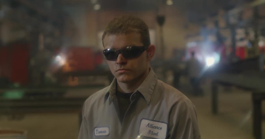 A man stands in a steel factory with sparks flying in the background