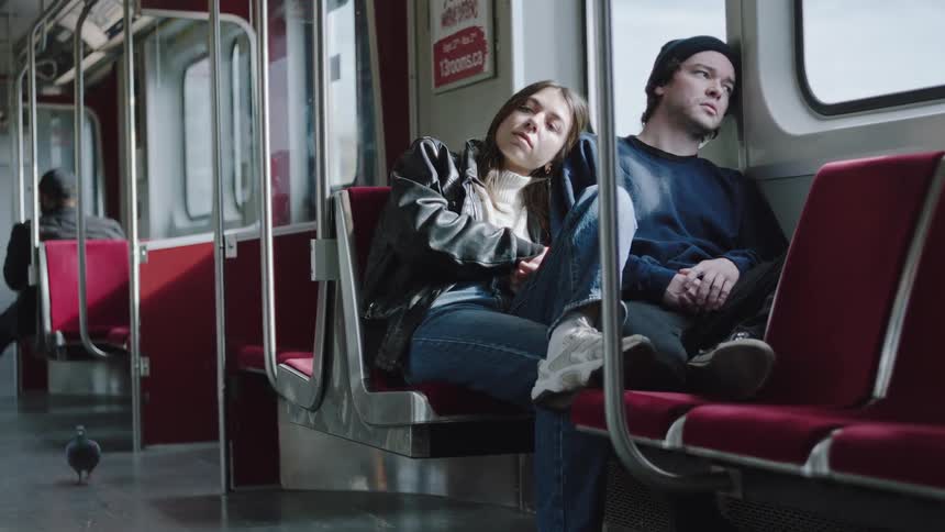Young adult couple sitting on train seats and leaning over each other with a contemplative facial expression with a pigeon running around them indoors during day time