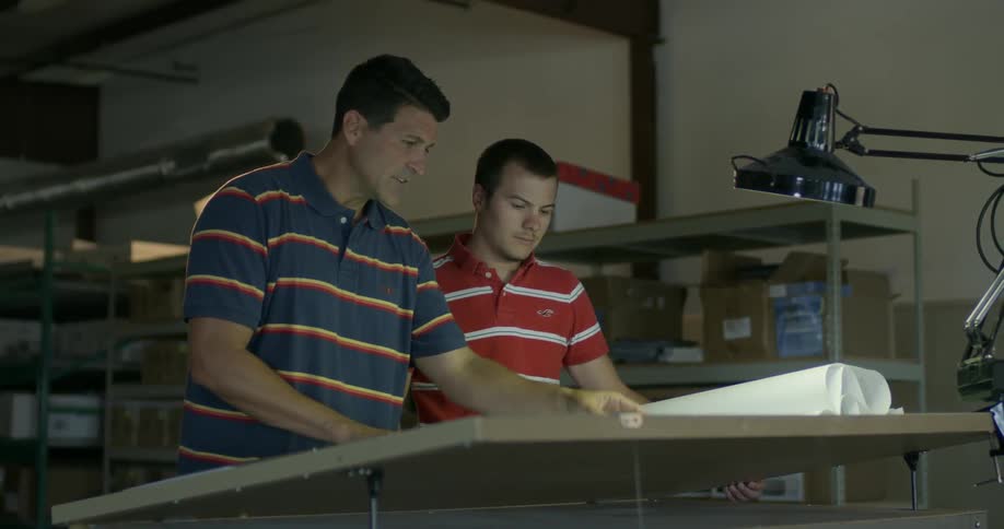 Two men look over a large sheet of paper in a workshop