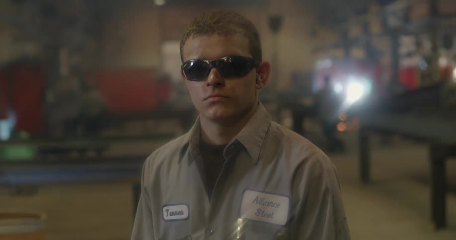A man wearing sunglasses stands in a workshop with men working behind him