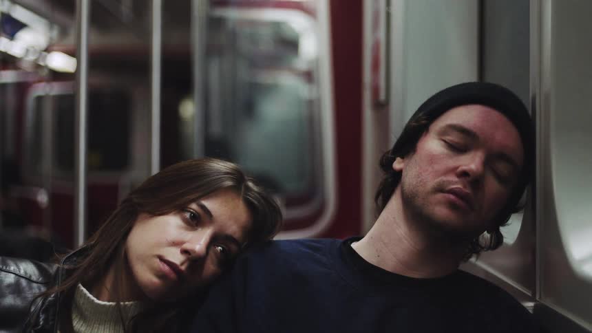 The video clip focuses on a young couple riding what appears to be a subway or train the man wearing a black beanie is asleep and leaning his head against the wall while the woman rests her head on his shoulder looking directly toward the camera with