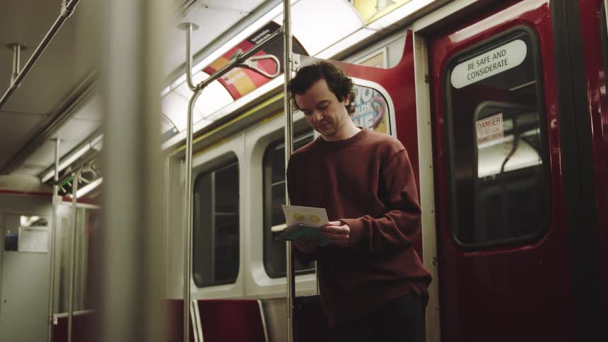 A smiling man stands reading a small card on a subway train