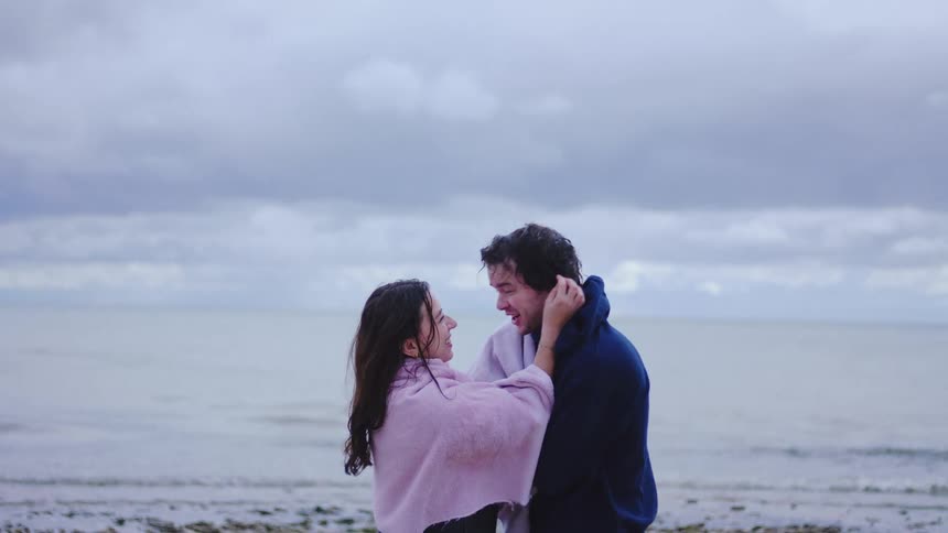 Couple standing on the beach with blankets over their shoulders in front of a cloudy sea