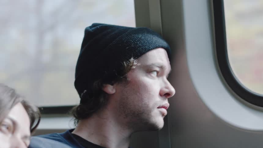This video clip shows a closeup of a young man riding a train or bus looking out the windowthe man is wearing a black knit beanie and has stubble and longish dark hair visible under the hathe is looking contemplatively out the window and anothe