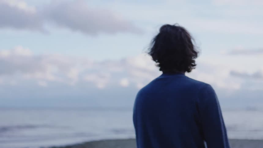 A man in a blue sweater stands up from a driftwood log to skip a stone across the water