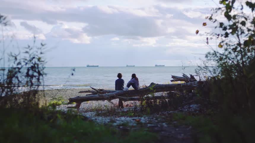 A man leans against a driftwood log before standing up to throw a rock into the ocean while a woman sits beside him looking out at the water