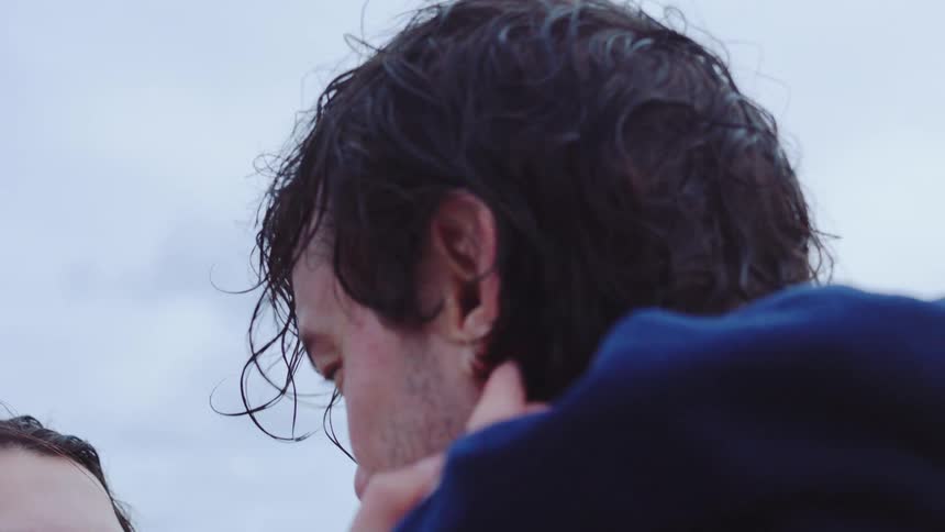 A closeup shot captures a young woman with a genuine emotional expression looking up at a man with wet hair who is embracing her outdoors under an overcast sky