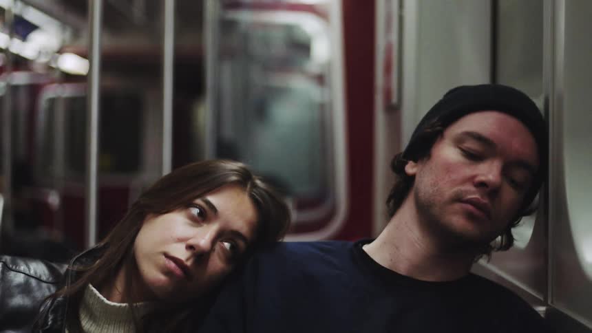 Two people seated side by side on a subway rest their heads against each other during the ride