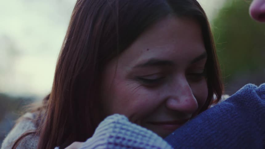 A closeup shot of a young womans face her eyes closing as she smiles softly and rests her head on a mans shoulder while embracing him outdoors in a moment of quiet affection