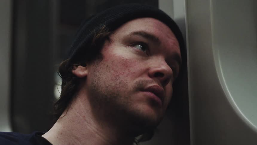 Young adult man wearing a black beanie sitting on a train seat leaning his head against the window indoors