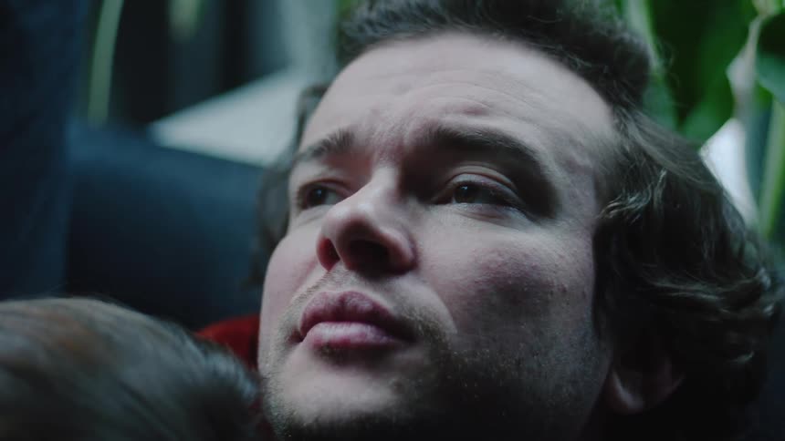Young adult man sitting down with a contemplative facial expression and a young adult woman leaning her head against his shoulder indoors