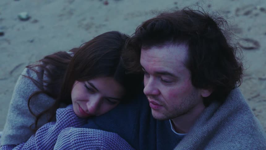 A couple cuddles closely on a beach the woman smiling softly as they share a quiet affectionate moment