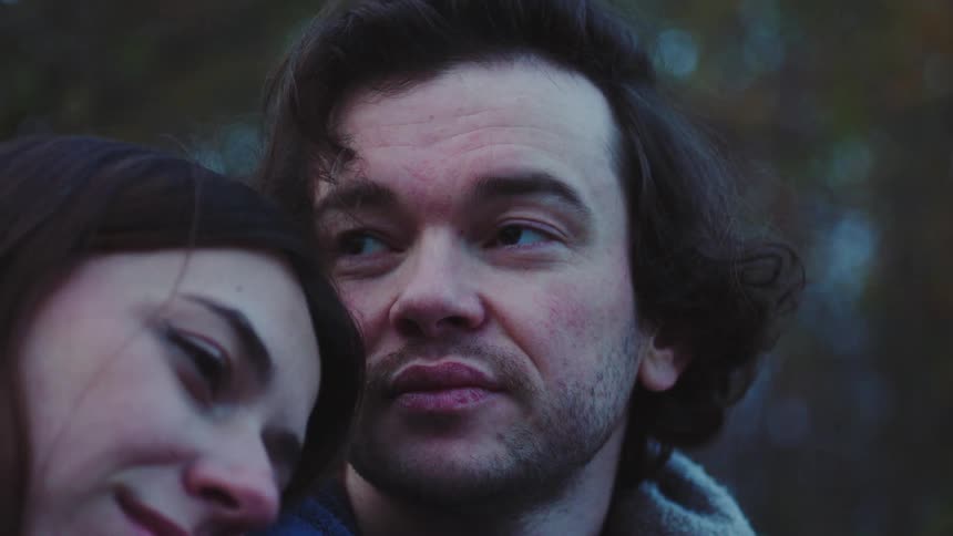 A man with curly hair gazes tenderly at a woman resting her head on his shoulder sharing a quiet intimate moment
