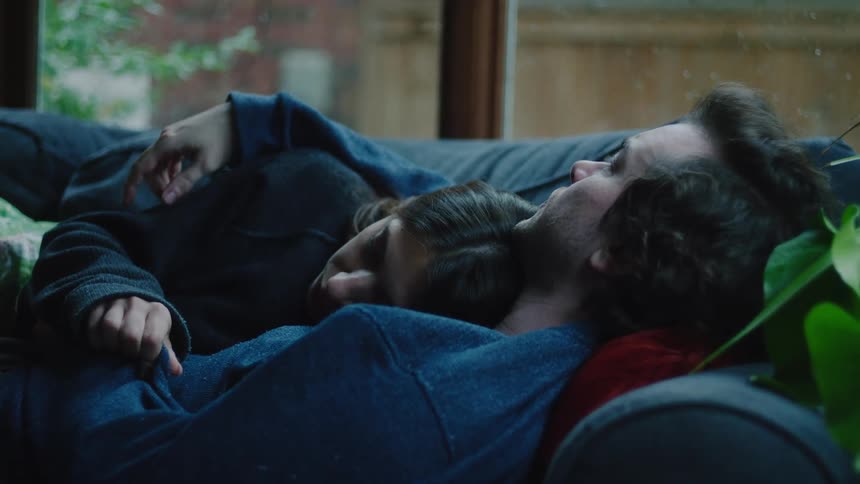 A man and woman lie together on a couch gazing at each other with affection in a cozy intimate moment