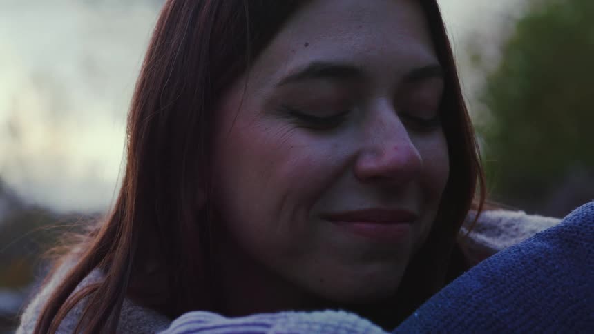 The woman with long dark hair is initially shown in profile looking off into the distance against a blurred overcast outdoor backgroundshe closes her eyes and gently rests her cheek against a blue knitted material suggesting a moment of peace or c