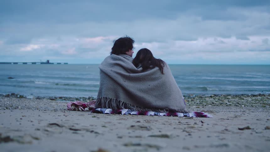 Couple sitting on the beach wrapped in a blanket as waves roll in to shore