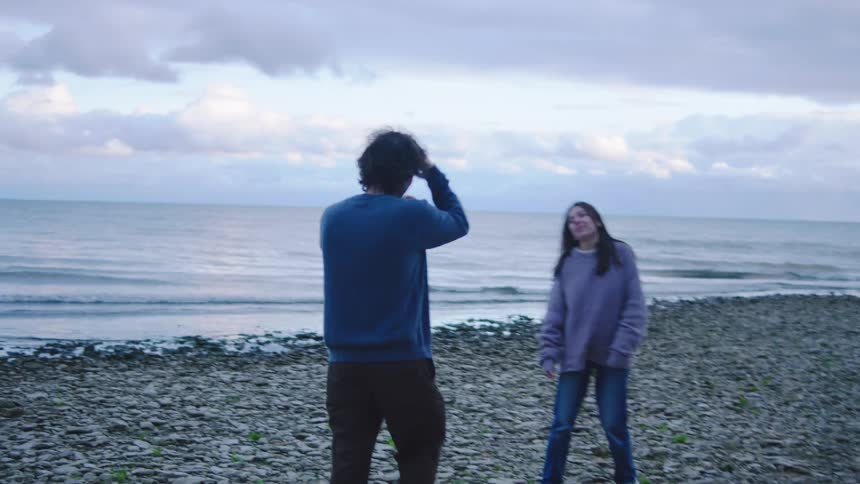A man is taking pictures of a woman who is standing on a beach and they are both wearing coats
