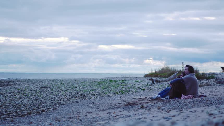 A man and a woman are wearing coats while sitting on the sand of an empty beach