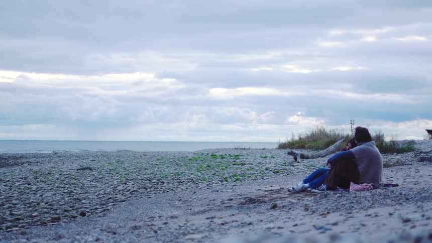 Young adult man and young adult woman covered in a blanket talking and hugging next to a beach outdoors