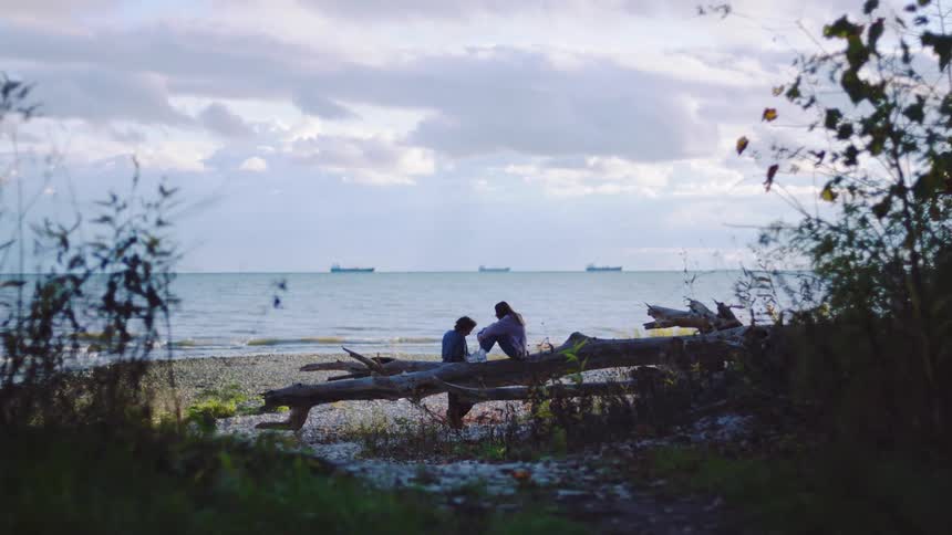 A wide steady shot of a couple sitting on a large piece of driftwood on a rocky shore framed by dark foliage with the vast ocean and a cloudy sky in the background