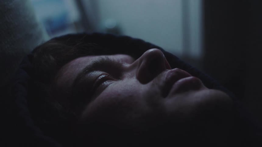 An extreme closeup of a mans face framed in shadow showing his eyes opening and looking up in a dark room