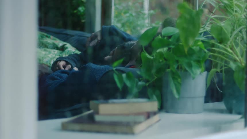 Young adult man and young adult woman cuddling on a sofa in a living room seen from outside the window during day time with rain pouring down