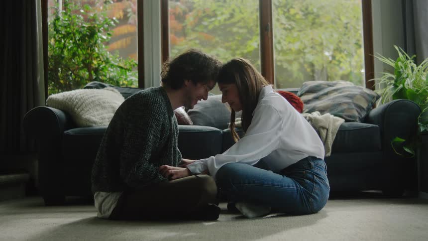 Young adult couple sitting down on the floor of a living room touching their foreheads and holding hands while they talk indoors during day time
