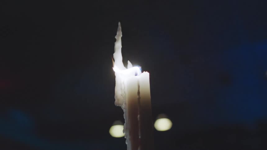 An extreme closeup of a thick white candle with melted wax dripping down the side showing the bright flickering flame against a dark background with small blurred lights