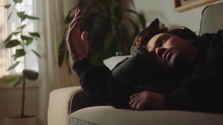 Young adult man lying on a couch looking at his own hand with a contemplative facial expression indoors during day time