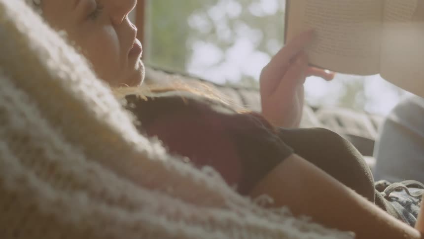 A woman is comfortably nestled under a knitted blanket on a couch relaxing and reading a book while talking to a man sittin on the floor all illuminated by bright natural light from a nearby window