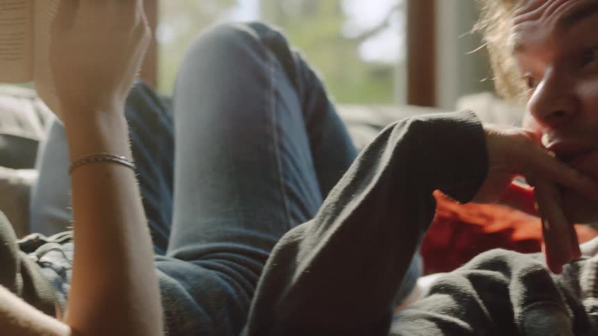 A man sits on a couch looking over at a woman who is lying down beside him reading a book
