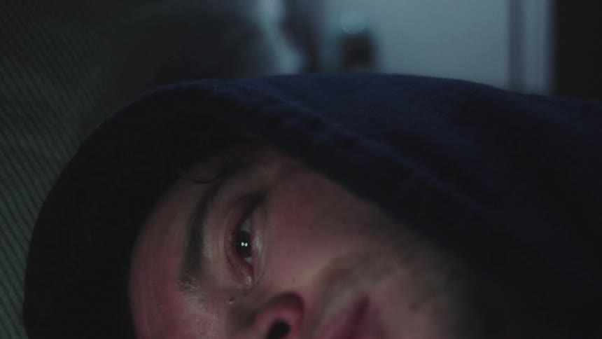 A young man wearing a dark hoodie lies with a distressed expression slowly opening his redrimmed eyes to look upward before shifting focus while the background remains softly blurred