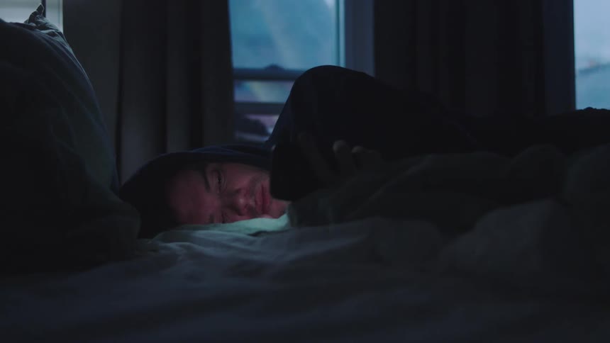 Young adult man wearing a black hoodie lying down in bed using a smartphone in a bedroom indoors
