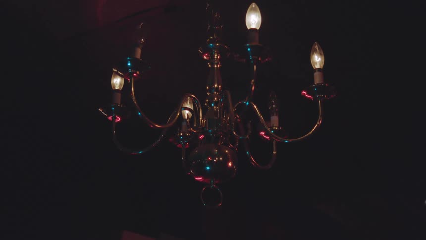 A classic brass chandelier featuring multiple arms and glowing candlestyle bulbs hangs centered in a dark room shimmering with reflections of its own light and a deep red glow in the back