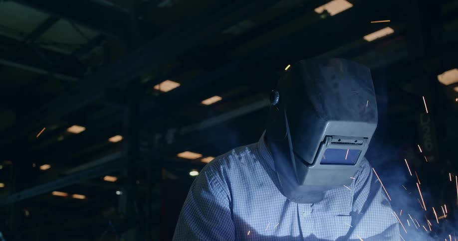 Person wearing a welding helmet while sparks fly in dark shop