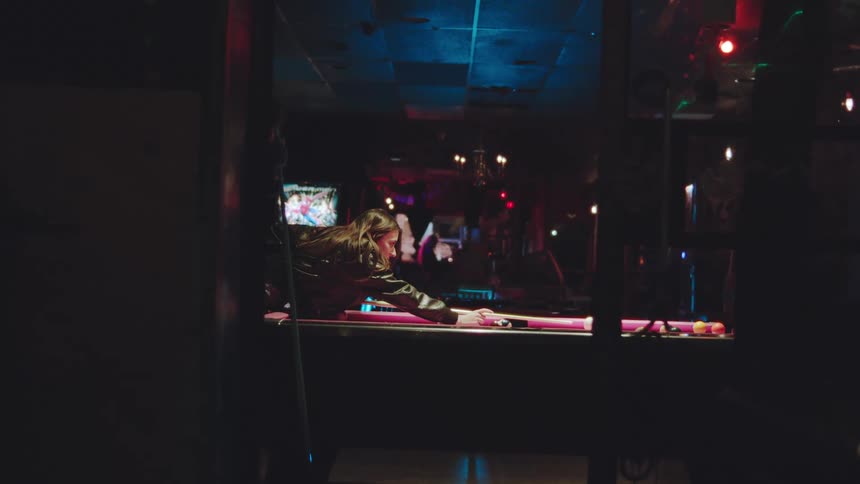 Woman leaning over red pool table aiming with cue stick in setting with colorful lights and tv in background
