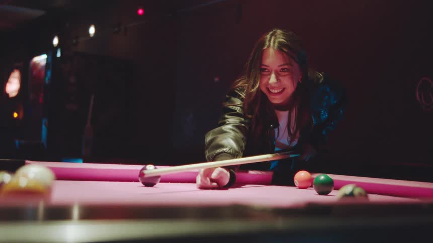 A woman wearing a leather jacket plays pool on a table with a distinctive pink felt in a dimly lit moody bar she smiles at the camera takes her shot and then looks excited and surprised by the outcome before the camera pans to another person in the da
